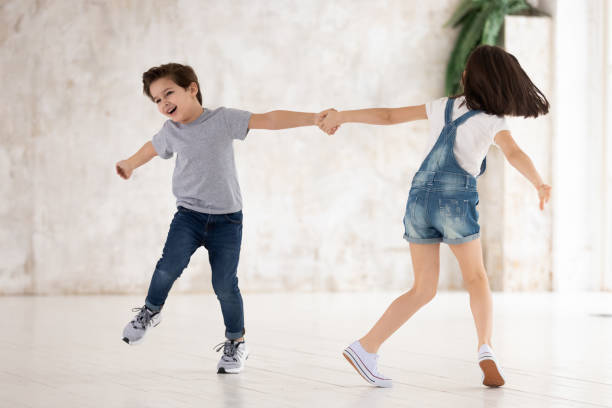 Excited funny little cute kids siblings playing active game at home, two happy small children boy and girl holding hands running spinning around laughing in living room in new modern empty apartment