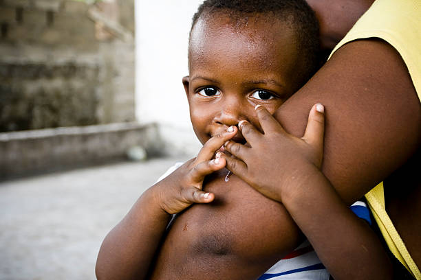 An African baby boy hugging his mothers arm while smiling at the camera.